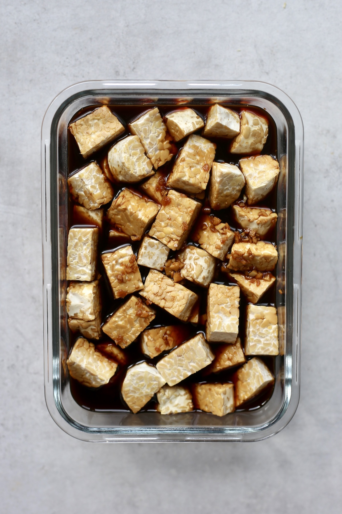 Cubes of tempeh soaking in a teriyaki marinade in a glass rectangle container. 