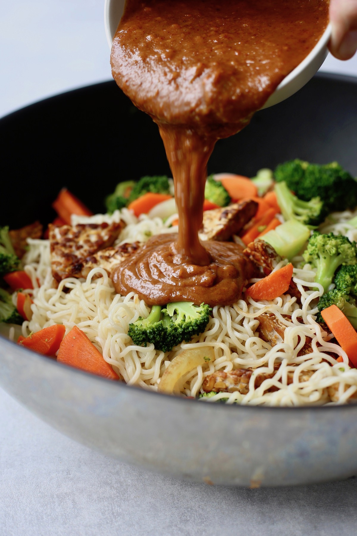 A thick almond butter sauce being poured over cooked noodles, veggies and tempeh in a large wok. 