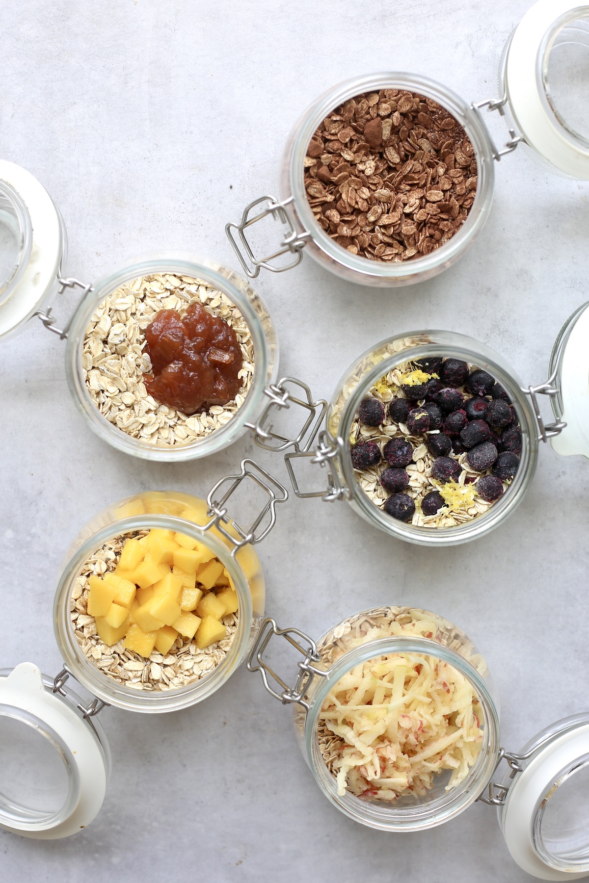 An overhead shot of chocolate, jelly, lemon blueberry, mango and apple overnight oats being prepared in wide mouth jars. 