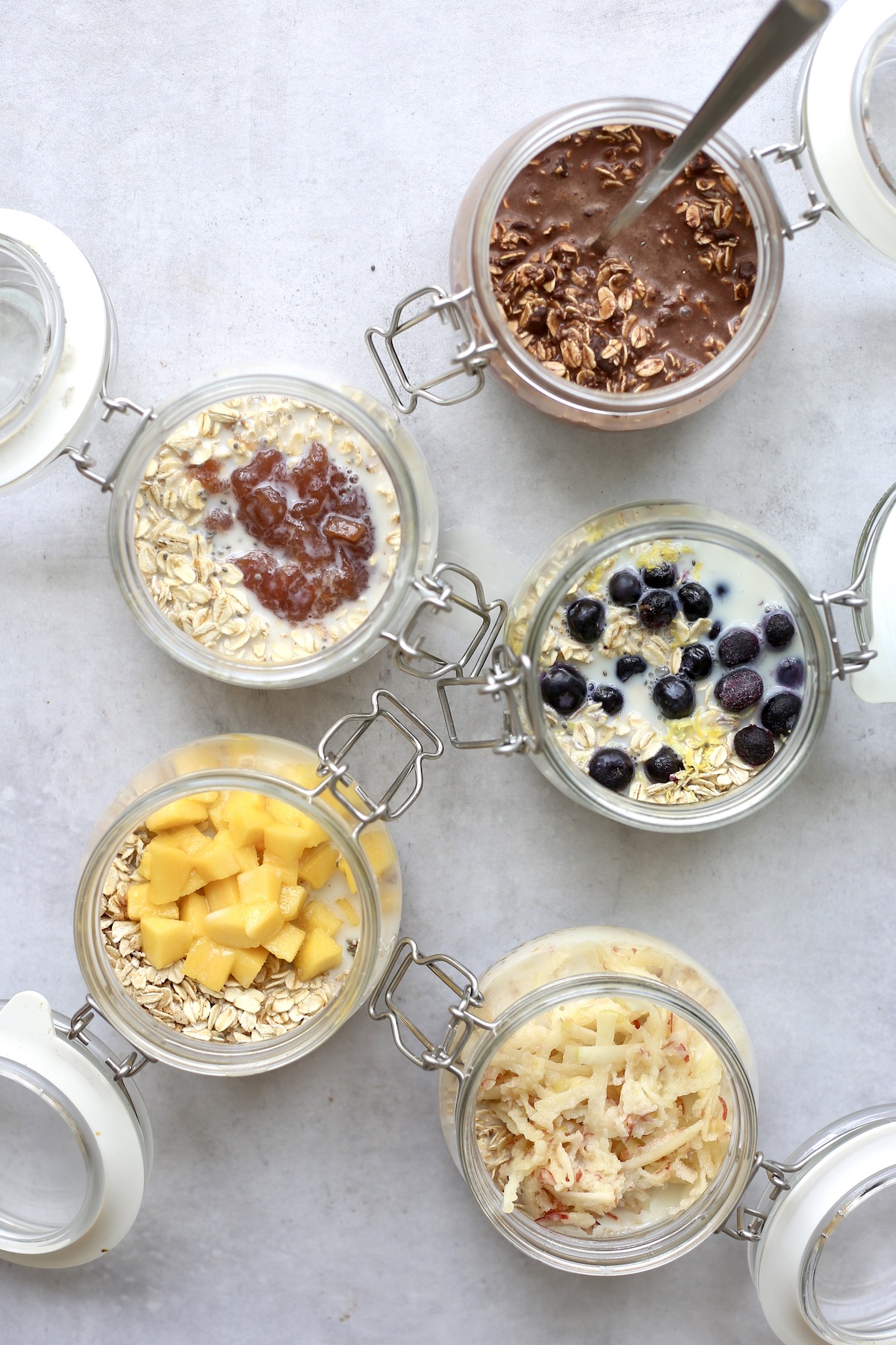 An overhead shot of 5 different flavors of overnight oats, one of which has a spoon the ingredients together.