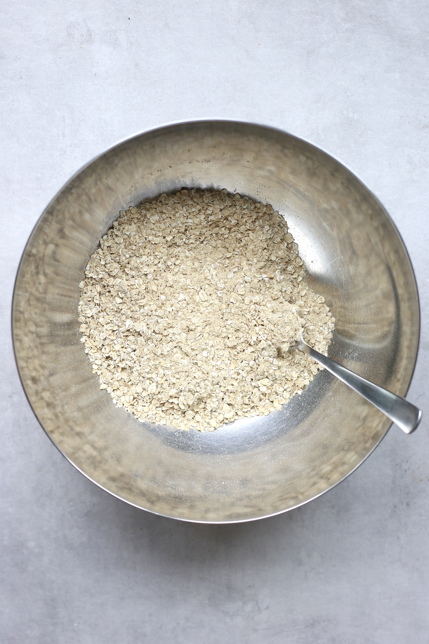 Quick oats, vegan protein powder, and ground flaxseed being stirred together in a large silver mixing bowl. 