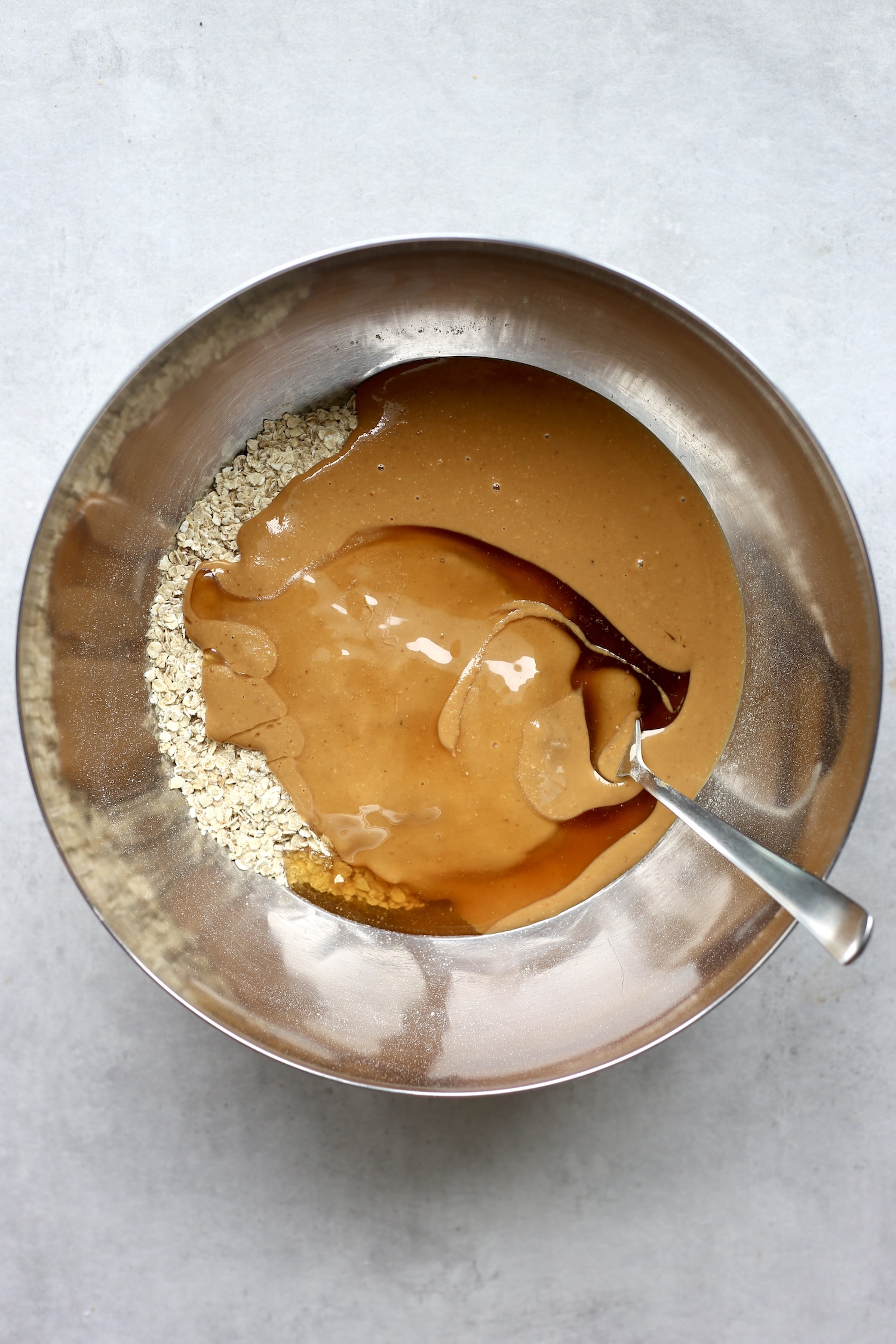 Peanut butter and honey being stirred into quick oats and vegan protein powder in a large silver mixing bowl. 