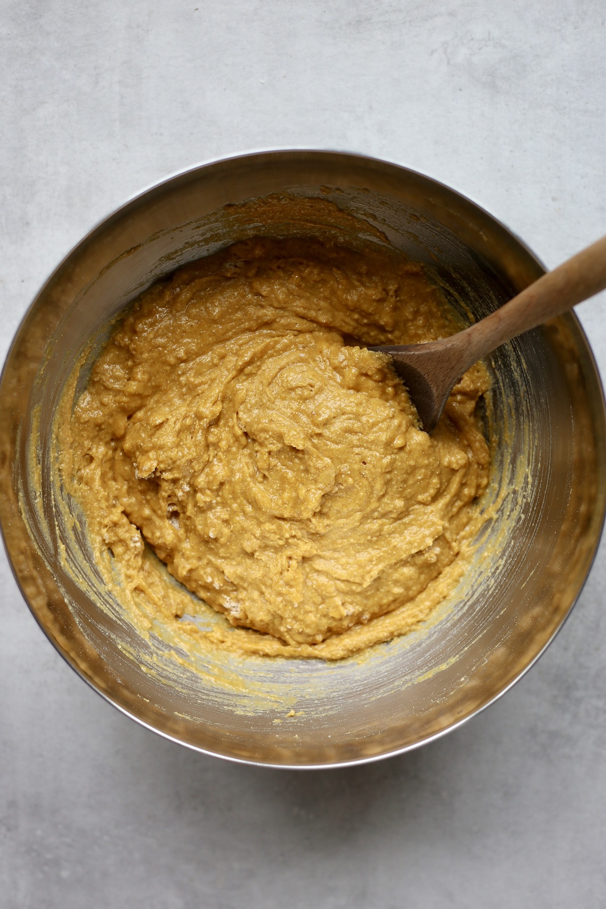 A wooden spoon stirring the batter for vegan pumpkin cornbread muffins in a large silver mixing bowl.