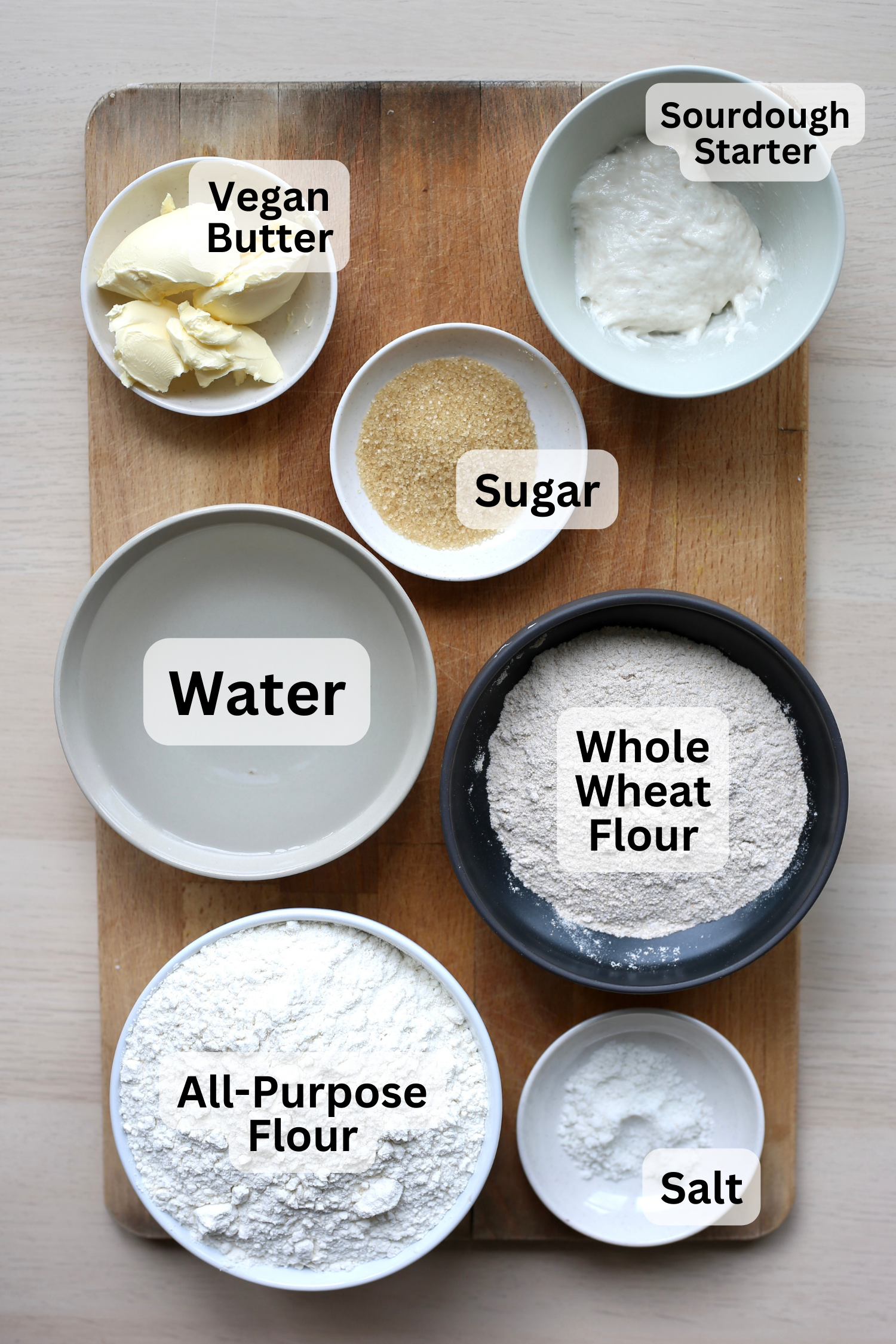 All of the ingredients for vegan sourdough bread measured out into bowls and laid out on a cutting board.