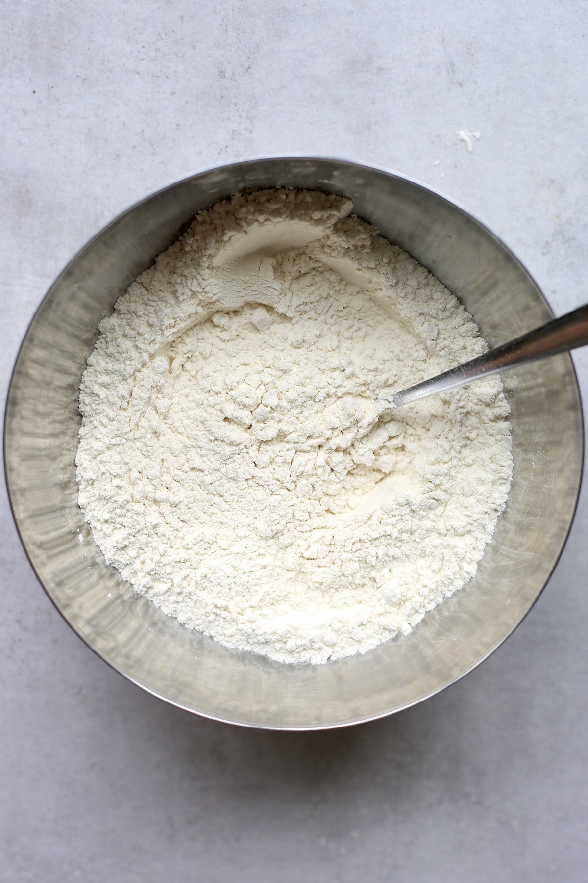 An overhead shot of flour, baking powder and salt being stirred together in a large silver mixing bowl with a silver spoon.