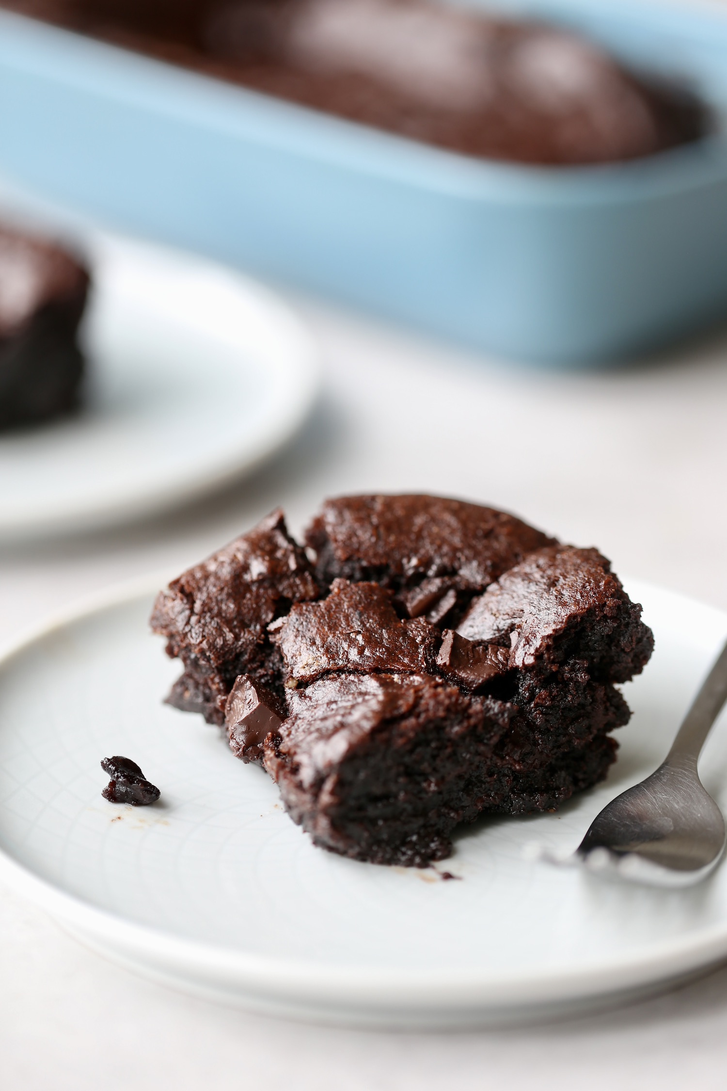 A gooey, thick tahini brownie on a white place with a silver fork beside it and more brownies in the background. 