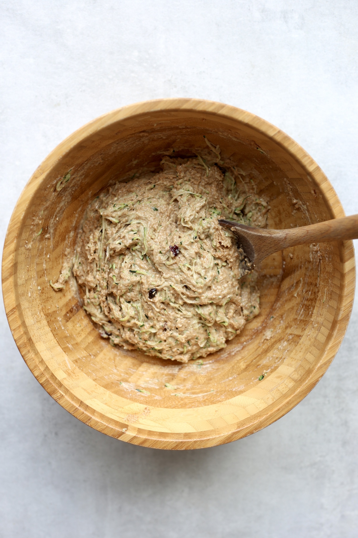 Shredded zucchini and raisins being stirred into muffin batter with a wooden spoon in a large wooden mixing bowl. 