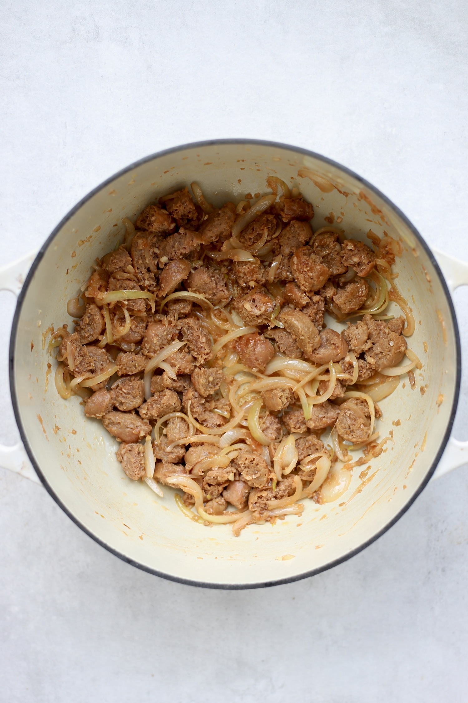 Vegan sausage and sliced onions browning in a large white pot. 