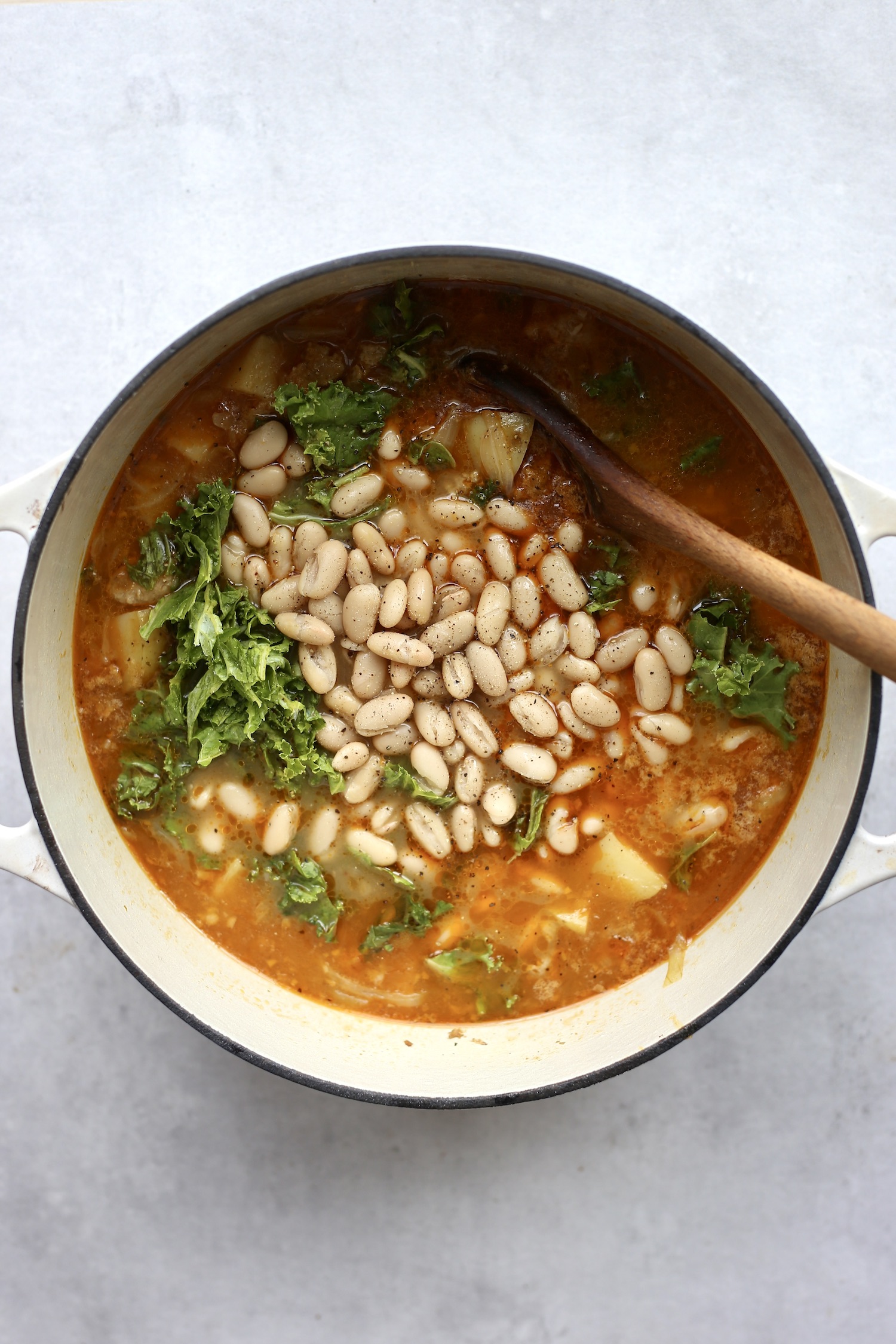 White beans, kale and black pepper being stirred into a sausage and potato soup.