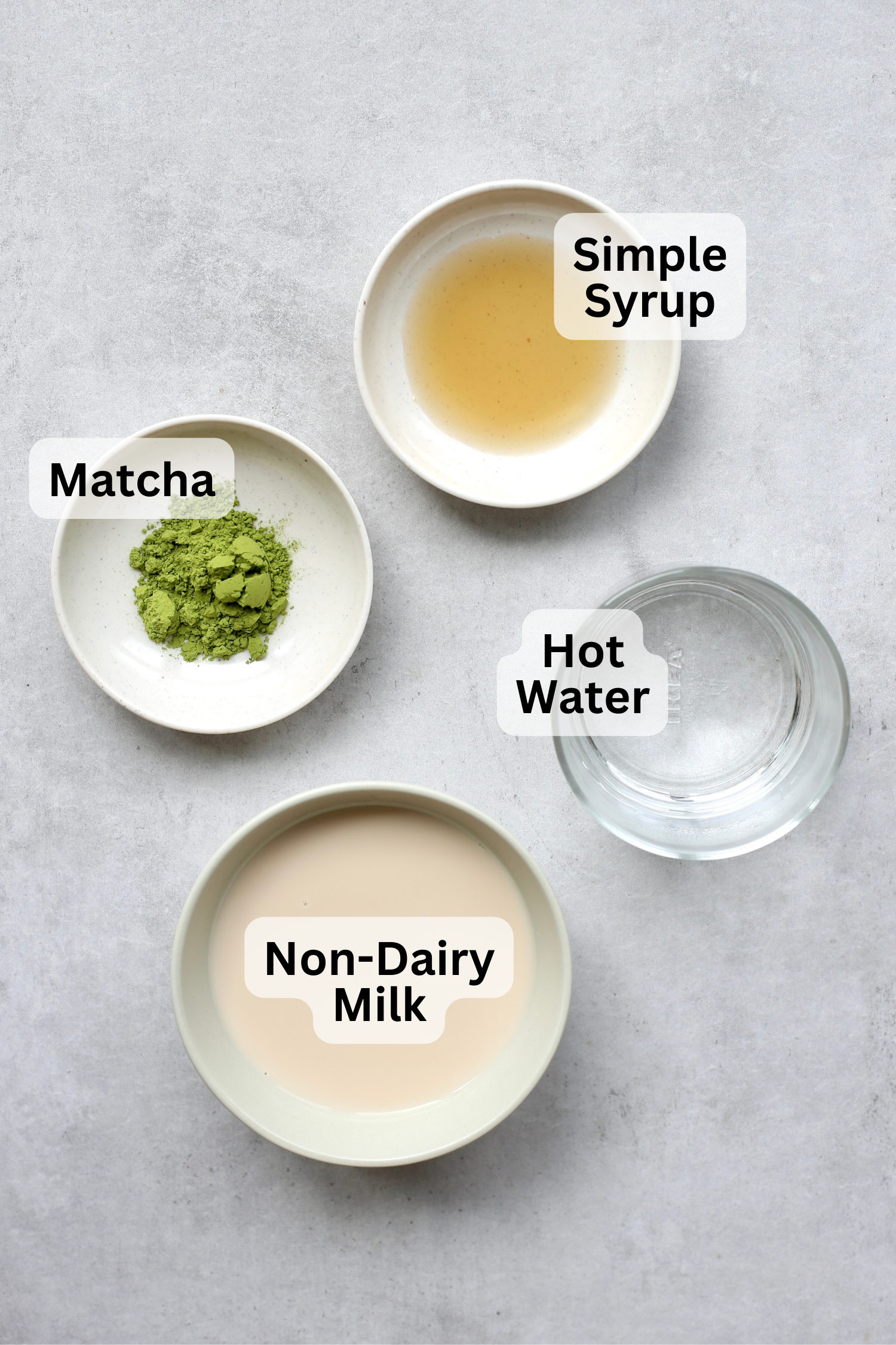 Simple syrup, matcha powder, hot water and non-dairy milk measured out into containers with labels overtop. 