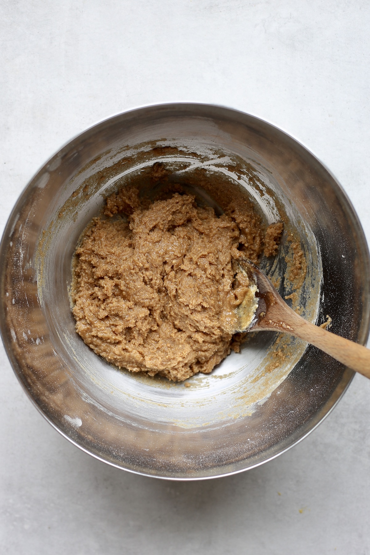 A wooden spoon stirring sticky peanut butter cookie dough in a large silver mixing bowl.