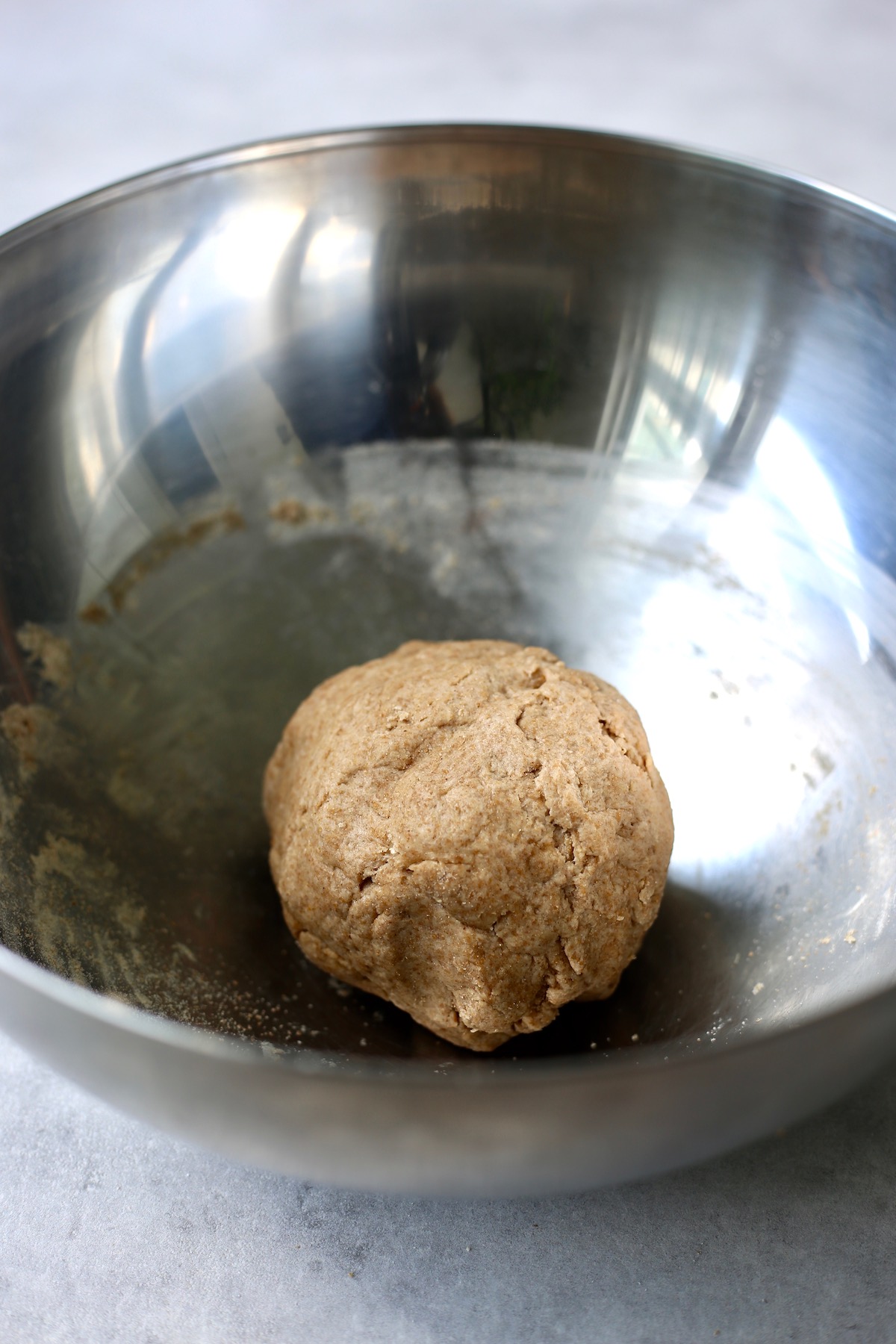 A ball of whole wheat pizza dough resting in a large silver mixing bowl.