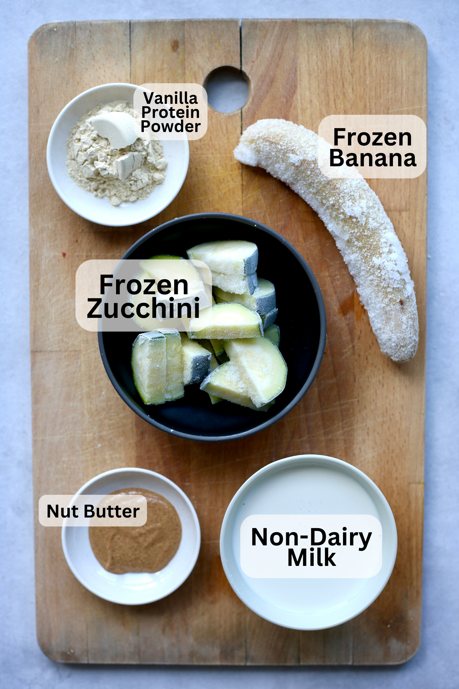 Vanilla protein powder, a frozen banana, frozen zucchini, milk and non-dairy milk measured out into bowls on a wooden cutting board. 